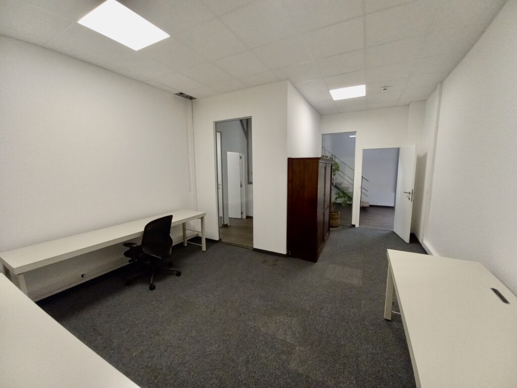 Een eenvoudige kantoorruimte met witte bureaus, één bureaustoel, grijs tapijt, een houten kast en een open deur naar een hal met trap.