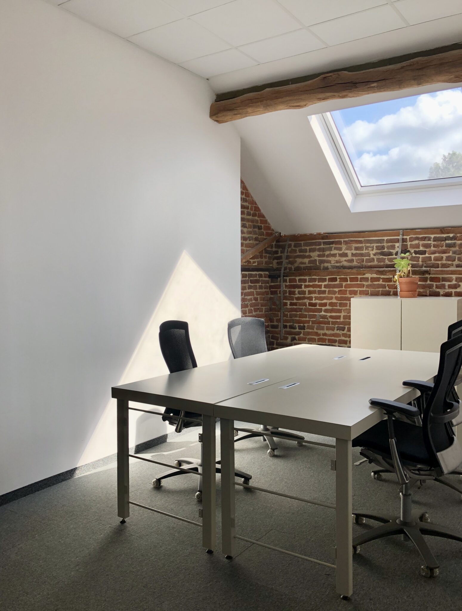 Een lichte kantoorruimte met vier bureaustoelen rond twee witte tafels, een dakraam, bakstenen muur en houten balken.