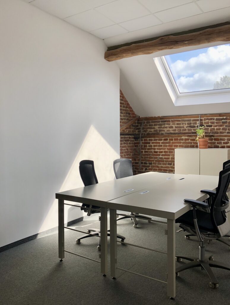 Een lichte kantoorruimte met vier bureaustoelen rond twee witte tafels, een dakraam, bakstenen muur en houten balken.