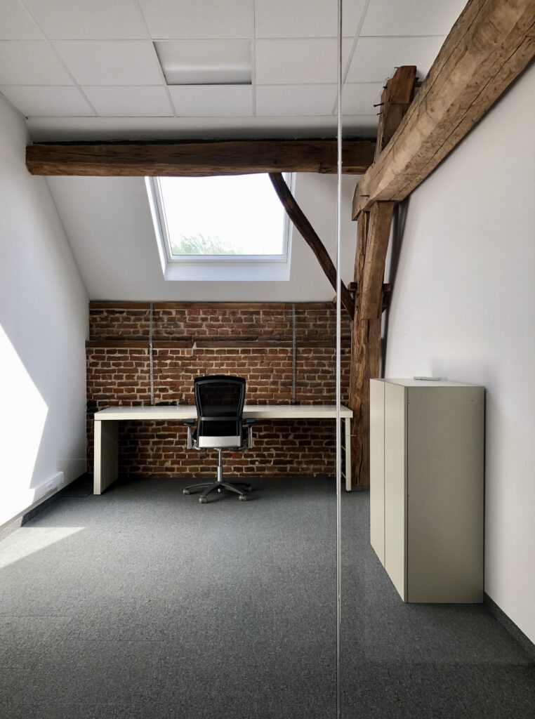 Een minimalistisch kantoor met bakstenen muur, wit bureau, bureaustoel, dakraam en zichtbare houten balken.
