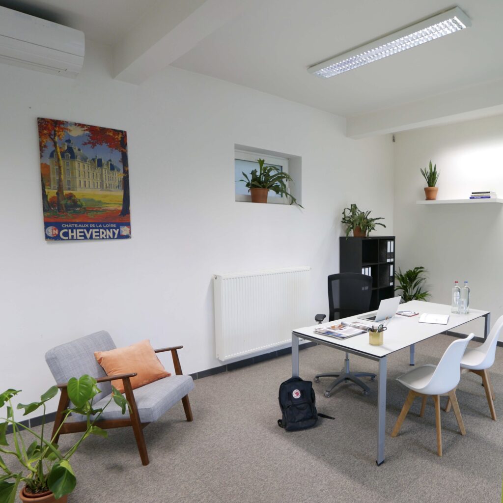 Een kantoorruimte met een bureau, stoelen, planten, een grijze fauteuil, een rugzak en een poster van Cheverny aan de muur.