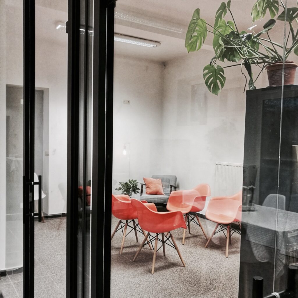 Een vergaderruimte met glazen wanden, vier oranje stoelen rond een lage tafel, een grijze fauteuil en meerdere kamerplanten.