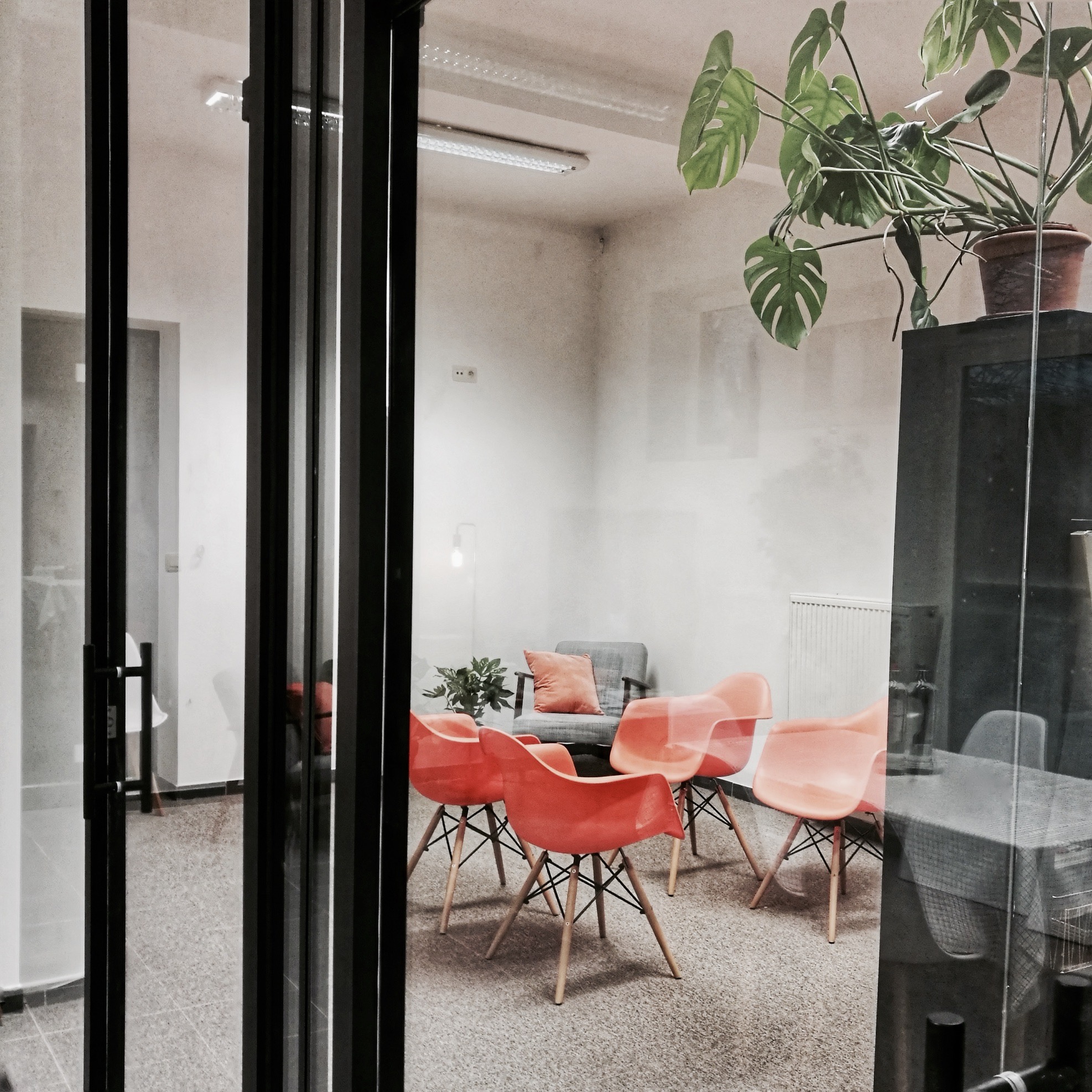 Een vergaderruimte met glazen wanden, vier oranje stoelen rond een lage tafel, een grijze fauteuil en meerdere kamerplanten.