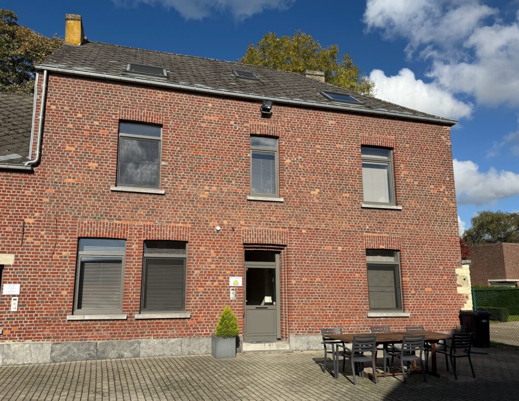Een hoge bakstenen woning met grijze voordeur, zes ramen met rolluiken en een buitenterras met tafel en stoelen op een verharde oprit. Bordje met logo van Klingspor.