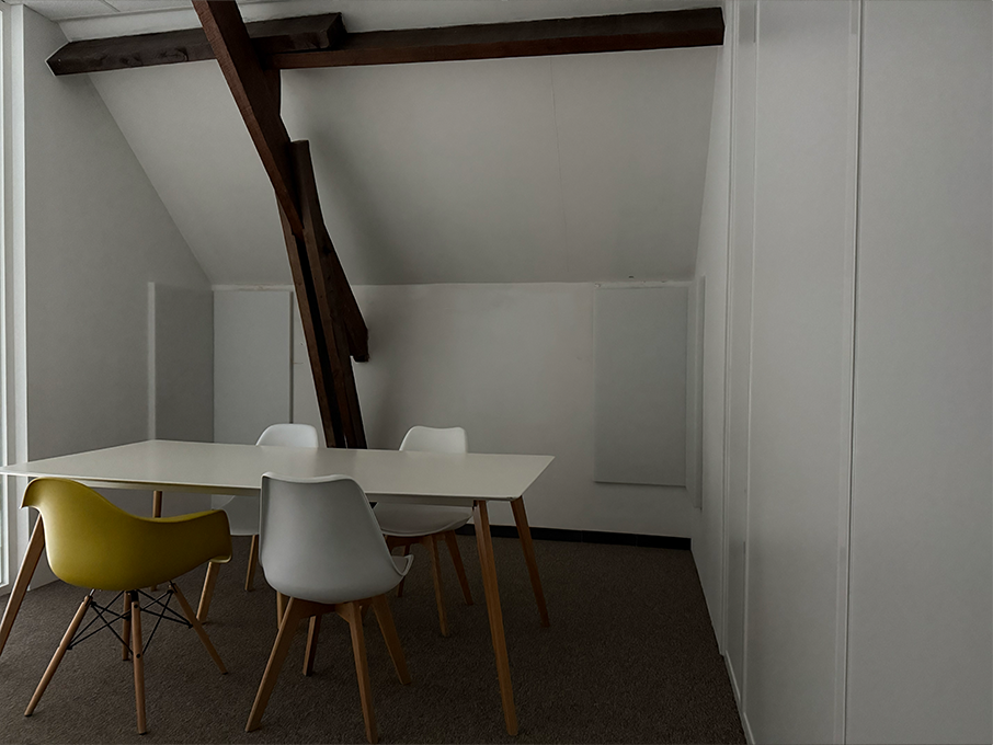 Een vergaderruimte met een witte tafel, vier stoelen en houten balken onder een schuin dak.