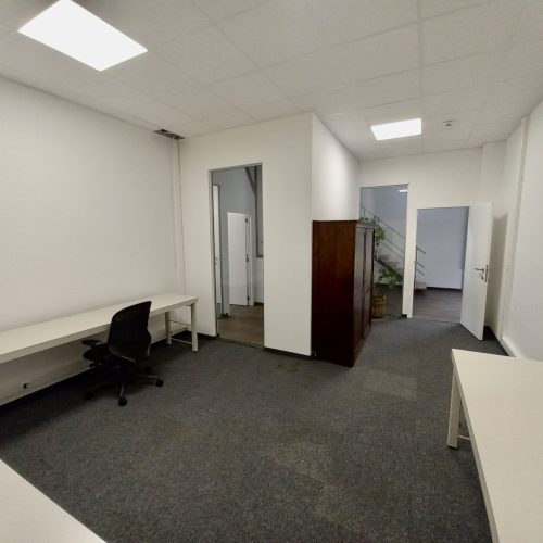 Een eenvoudige kantoorruimte met witte bureaus, één bureaustoel, grijs tapijt, een houten kast en een open deur naar een hal met trap.