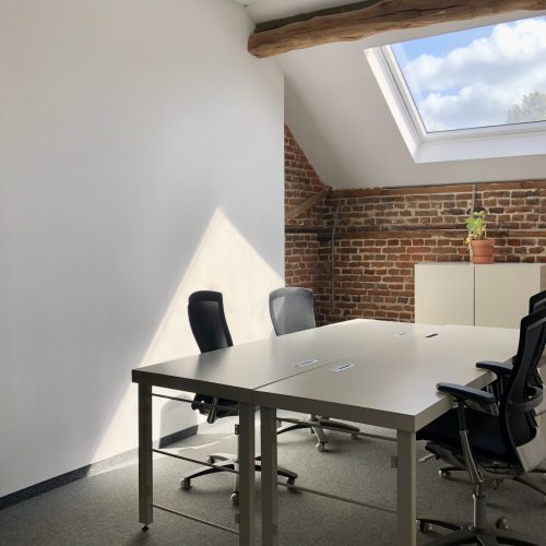 Een lichte kantoorruimte met vier bureaustoelen rond twee witte tafels, een dakraam, bakstenen muur en houten balken.