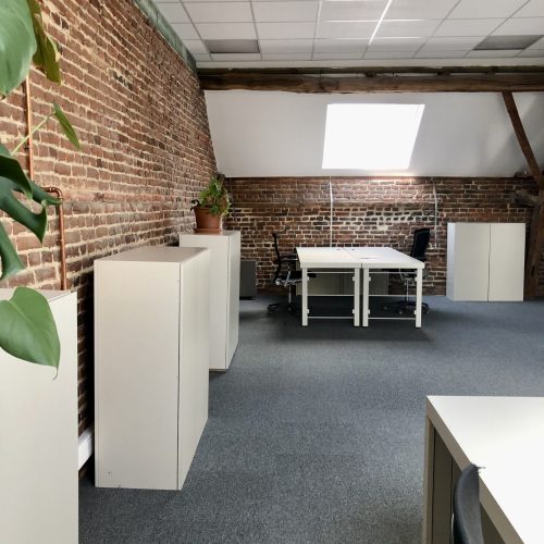 Rechterzicht van een kantoorruimte met zichtbare bakstenen muur, meerdere witte kasten, werkplekken met bureaustoelen, een dakraam en houten balken.