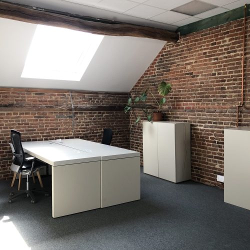 Linkerzicht van een ruime kantoorruimte met bakstenen muren, meerdere bureaus met bureaustoelen, een dakraam en enkele kasten met een kamerplant.