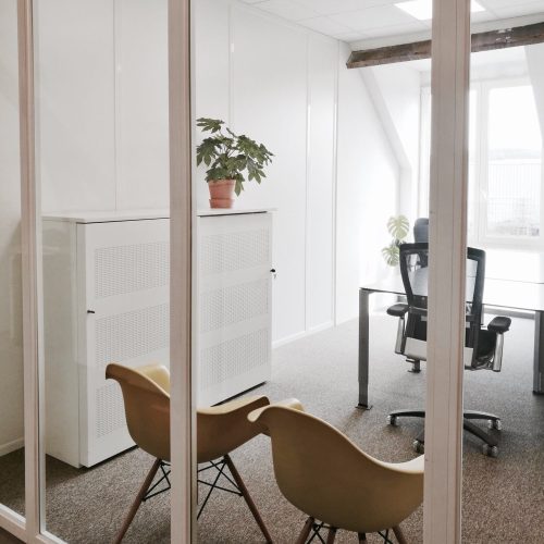 Een moderne kantoorruimte met glazen wanden, twee gele stoelen, een witte kast met plant, en een werkplek bij een groot raam.