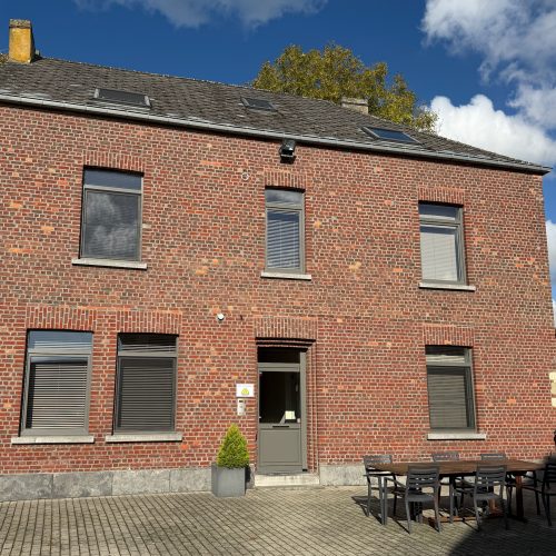 Een hoge bakstenen woning met grijze voordeur, zes ramen met rolluiken en een buitenterras met tafel en stoelen op een verharde oprit. Bordje met logo van Klingspor.