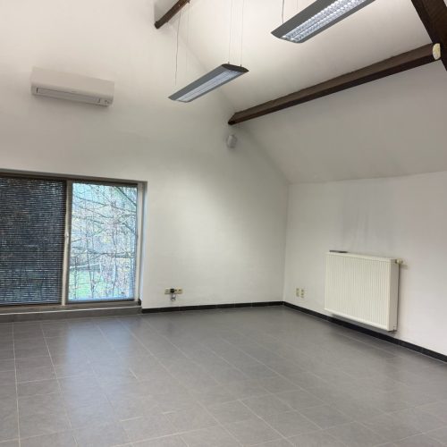 Lichte kantoorruimte met groot raam, radiator, airco en zichtbare houten balken.