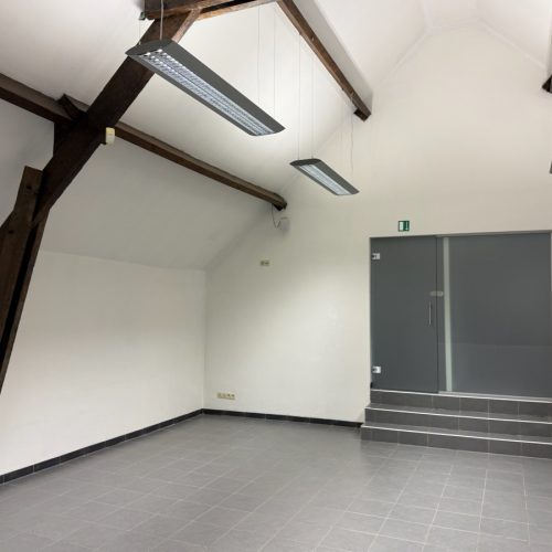 Lichte kantoorruimte met schuine wanden, zichtbare houten balken, grijze tegelvloer en glazen deur met trapje.