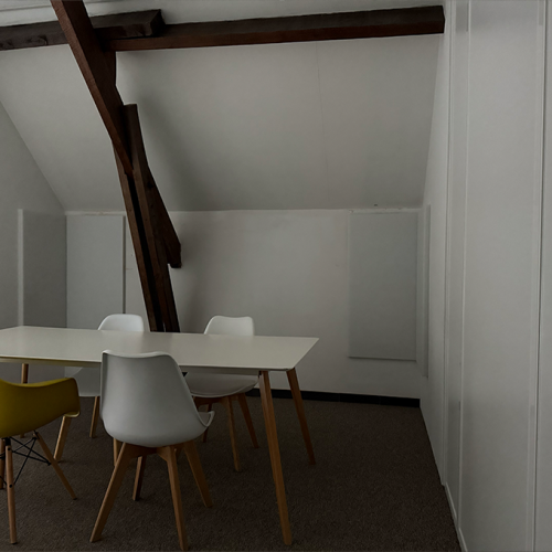 Een vergaderruimte met een witte tafel, vier stoelen en houten balken onder een schuin dak.