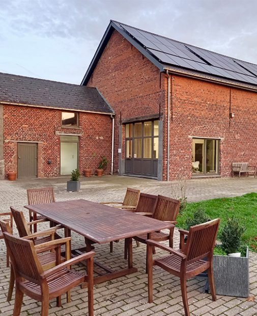 Een gerenoveerde bakstenen hoeve gebouw met zonnepanelen op het dak, grote ramen en een buitenterras met houten tuinmeubilair.